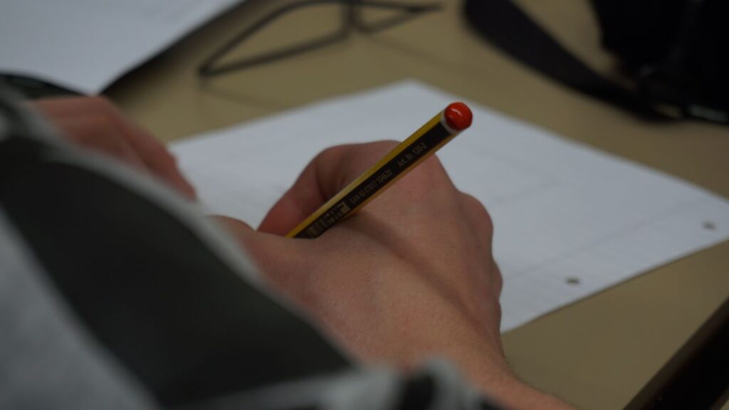 Hand holding a pencil writing on lined paper indoors, focus on detail.
