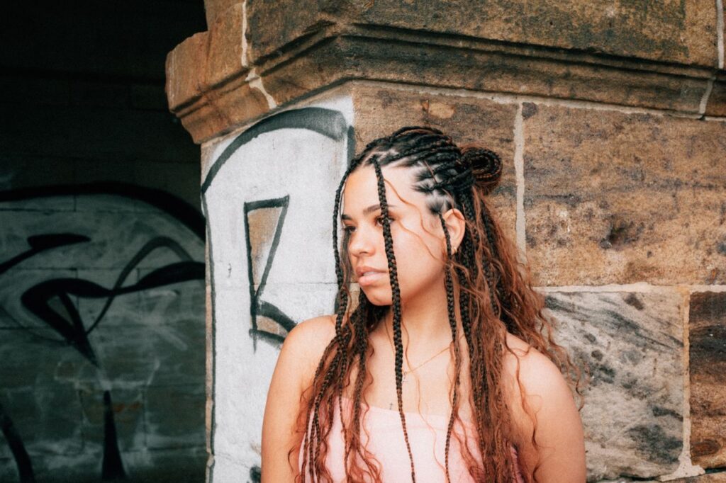 Portrait of a young woman with braids standing by a graffiti-covered wall, urban setting.
